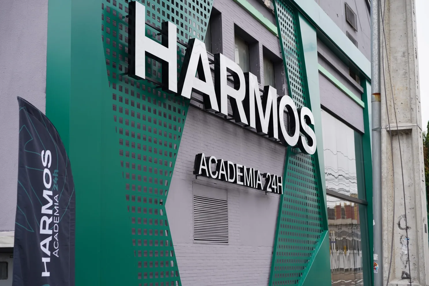 Fachada da Harmos Academia 24h, com painéis verdes e letreiro iluminado.